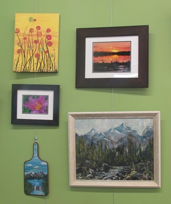 a closer view of the art wall described above. The paintings here are red flowers on a yellow background, a sunset in a brown frame, a close up of a pink flower, a mountain and lake landscape painted on a cutting board with a handle, and last but not least is a mountain landscape with beautiful trees and a raging river